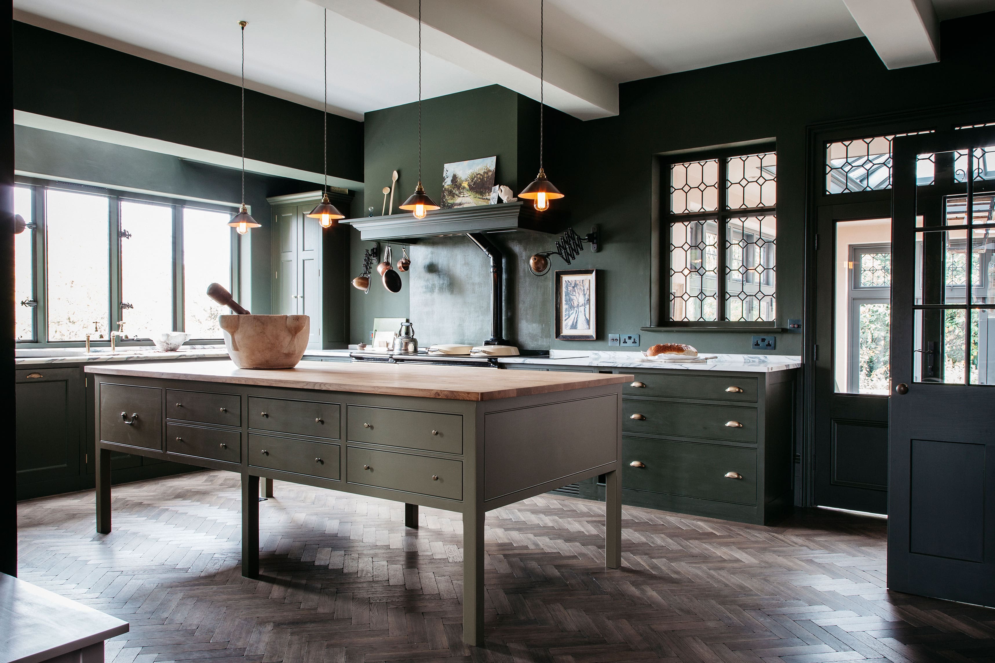  View of a spacious English kitchen featuring dark blue cabinetry, a wooden kitchen island, and a large range cooker with brass accents, designed by Plain English.