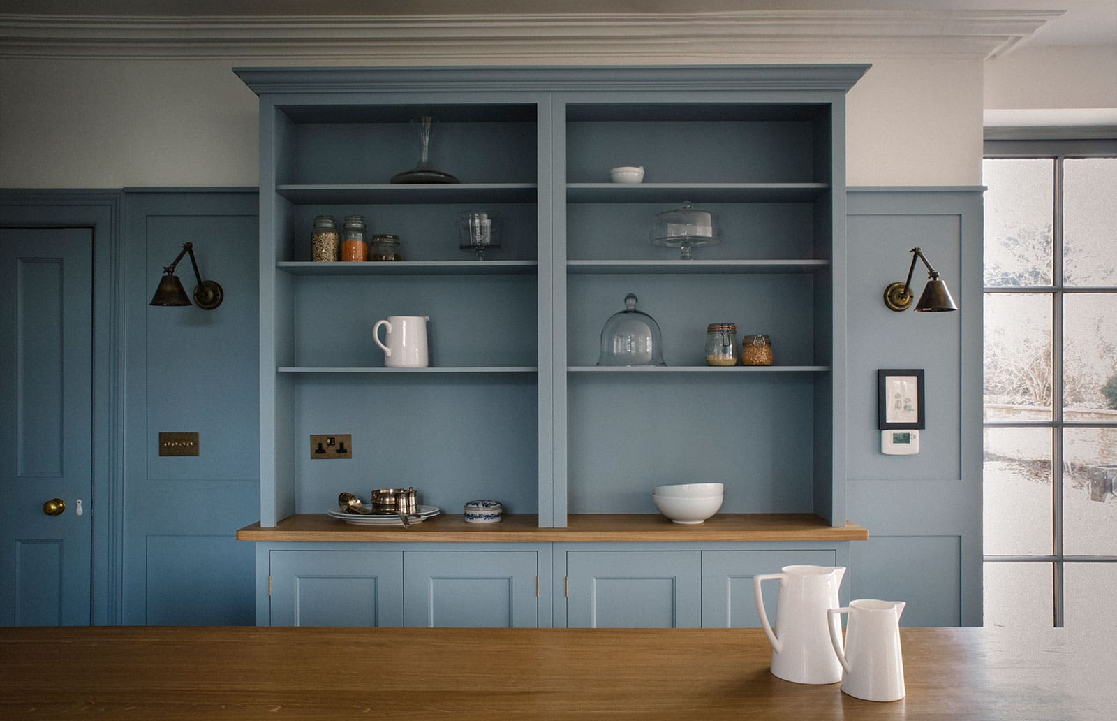  Blue kitchen shelves with jars, dishes, and white pitchers, set against wooden countertops and brass wall lights.