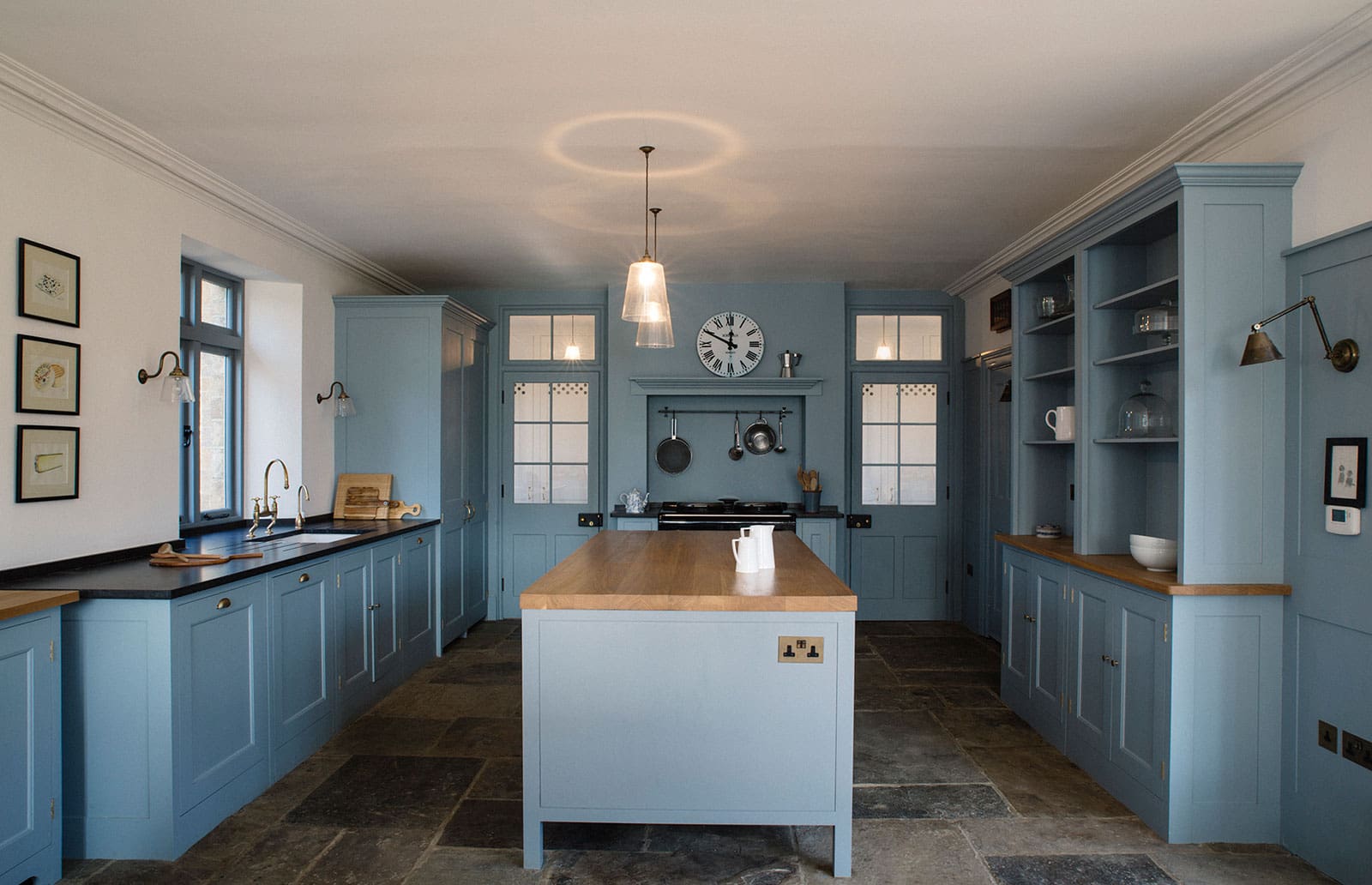  Kitchen with blue cabinetry, wooden-topped island, stone flooring, and a stove framed by a clock and hanging cookware.