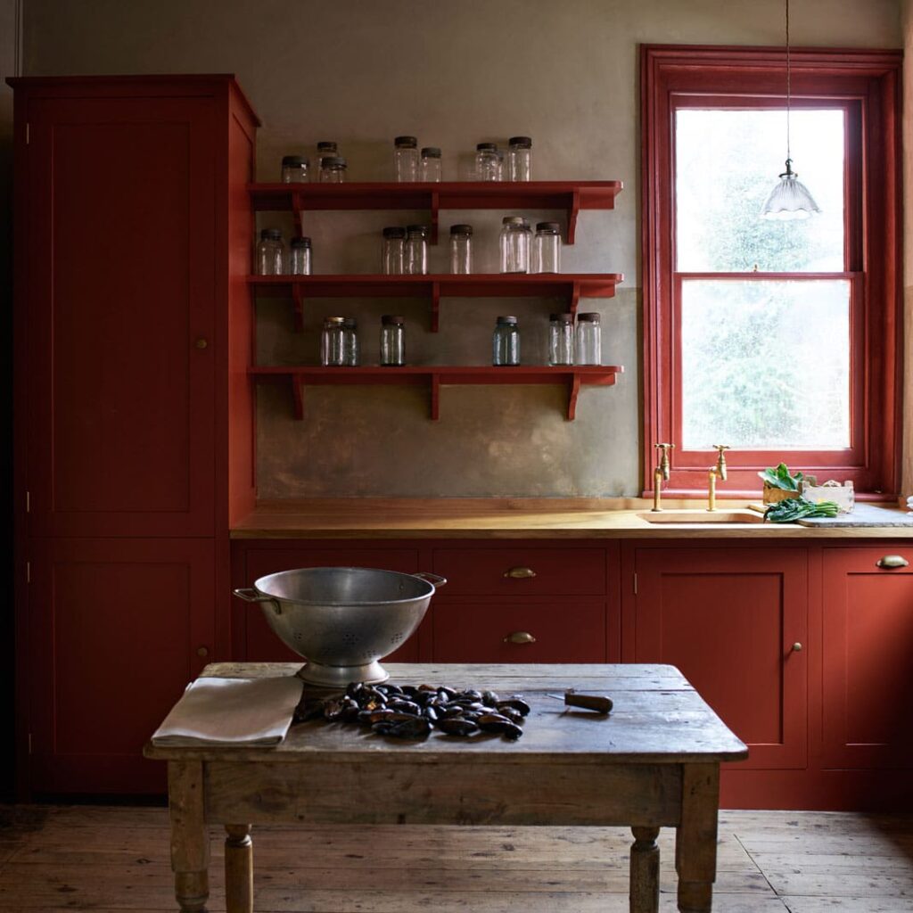 Rustic kitchen with red cabinetry, wooden table, and brass fixtures by the window.