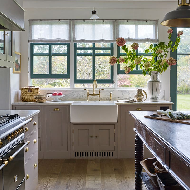 Kitchen with bespoke cabinetry in muted taupe with brass hardware, featuring a custom white farmhouse sink and marble countertop.