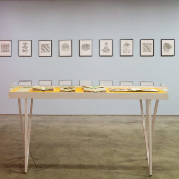 Wooden drawing table with a yellow top covered in books, with simple art hanging on the wall behind it.