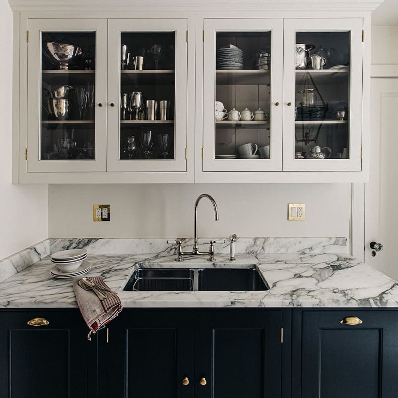 Kitchen sink with marble countertops and custom designed blue cabinets under the sink and white, glass door cabinets overhead.
