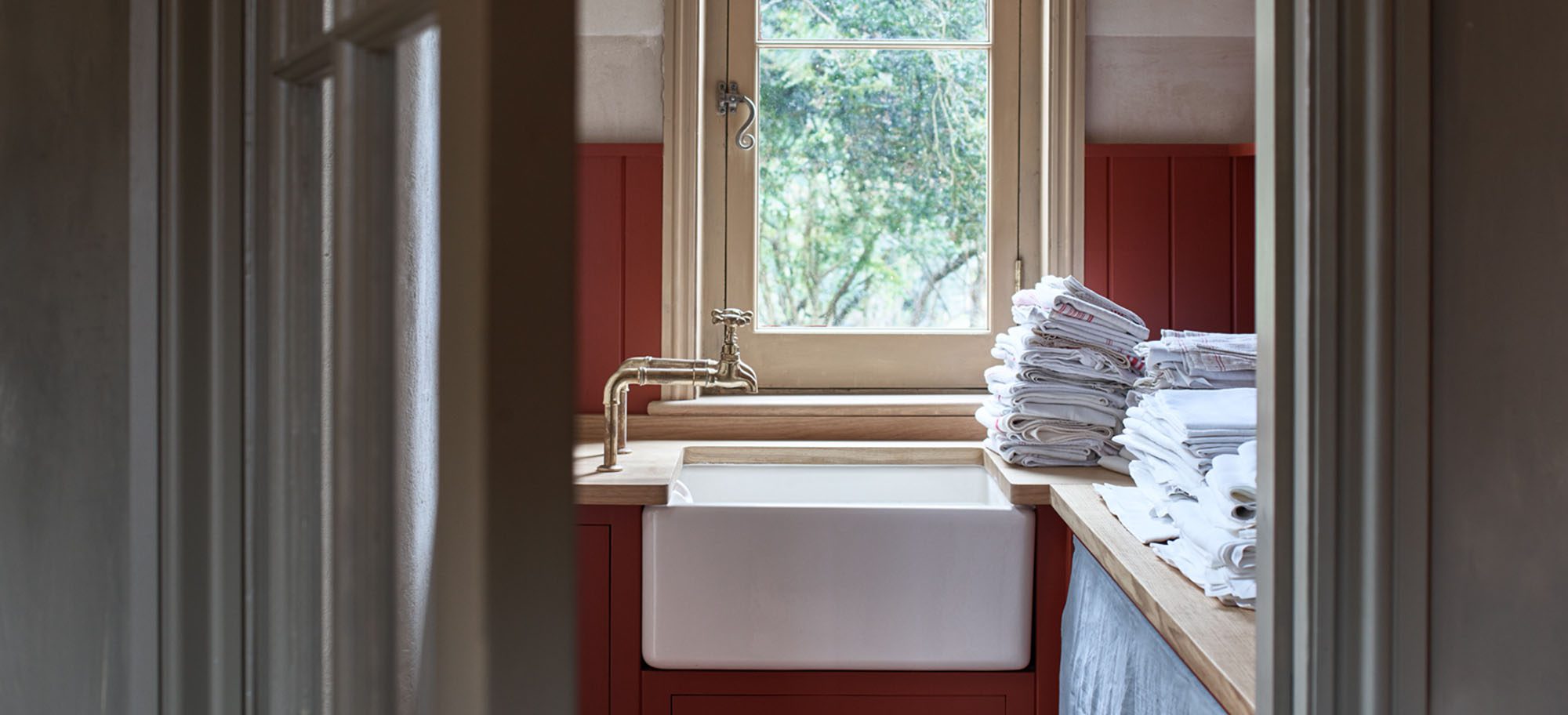 Bespoke laundry room design featuring red cabinetry and a farmhouse sink. A kitchen with a farmhouse sink, brass faucet, red cabinetry, and a stack of linens by a window overlooking greenery.