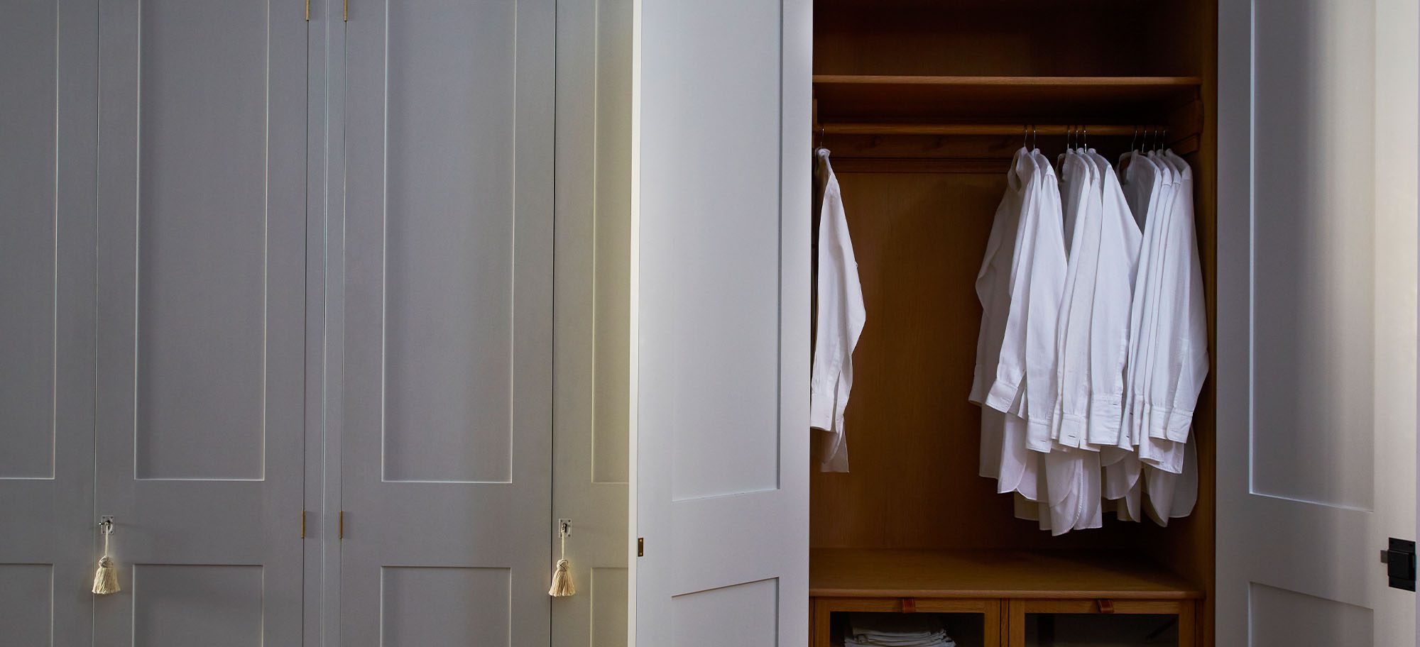 Custom bespoke wardrobes with white shirts hanging in a wooden interior. A partially open white wardrobe reveals neatly hung white shirts and wooden shelving inside.