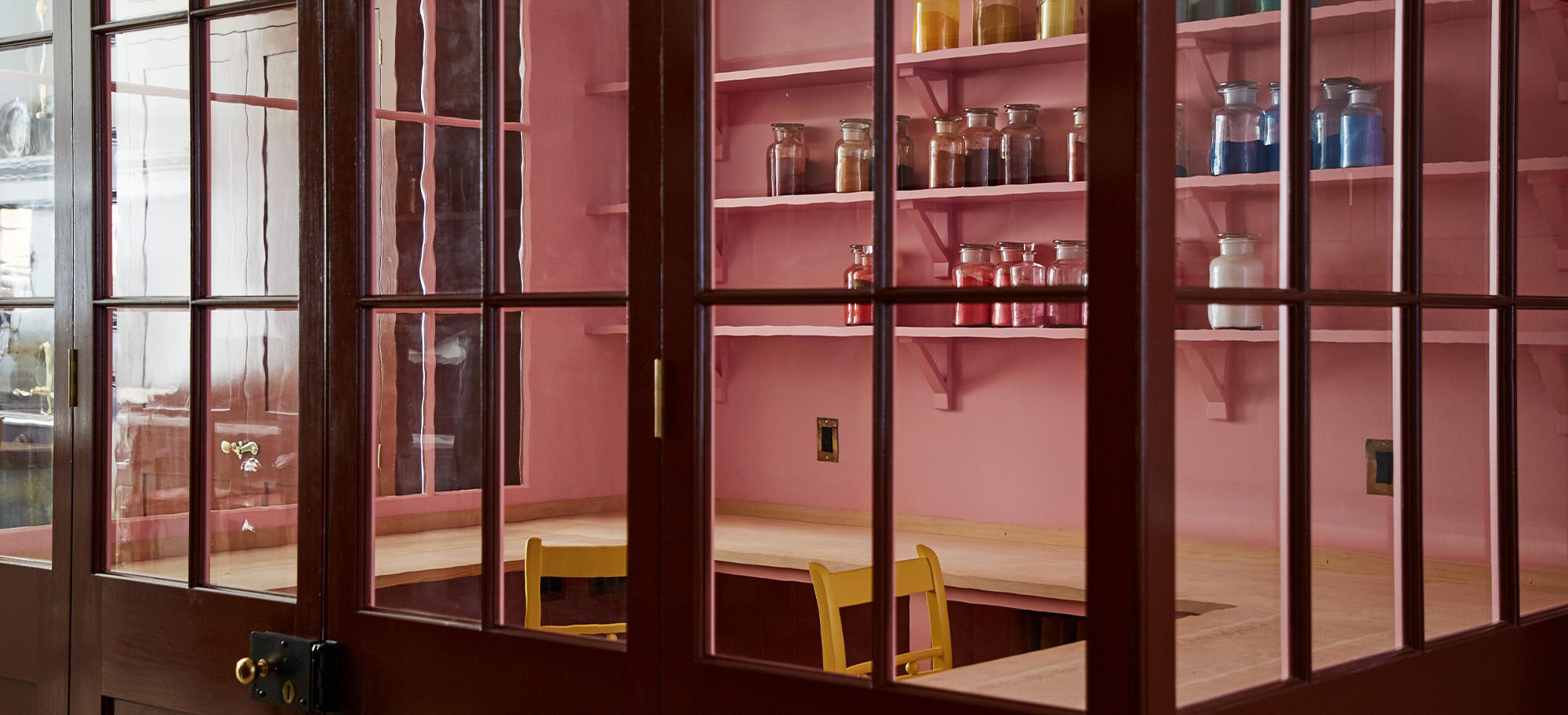 Pots and pans hanging along a custom designed kitchen, featuring neutral-coloured cabinets. Jars on shelves in light red room