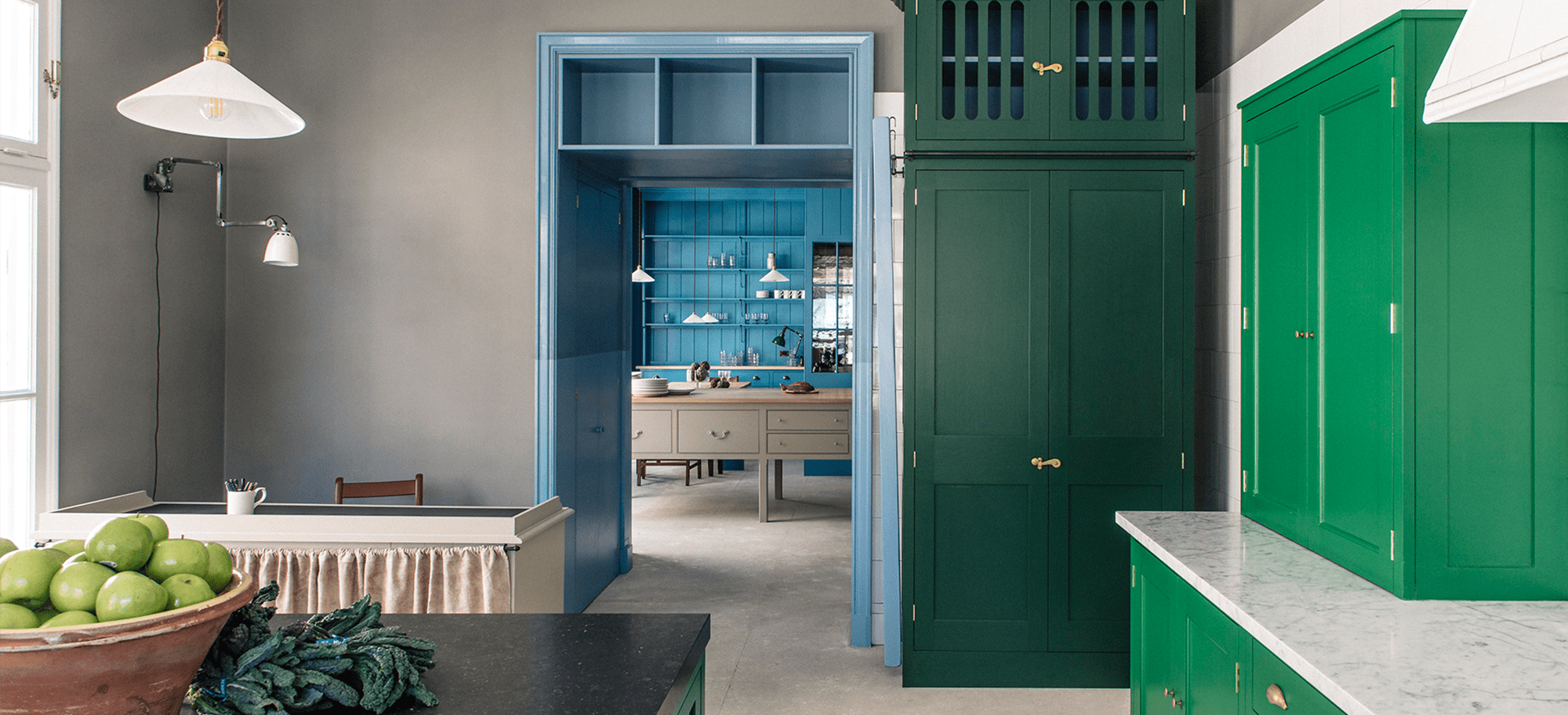 Bespoke Kitchen Cabinets Bespoke kitchen with deep green cabinetry and a marble countertop leads to a pantry featuring light blue paneling and a wooden table, creating a harmonious blend of colors and classic design.