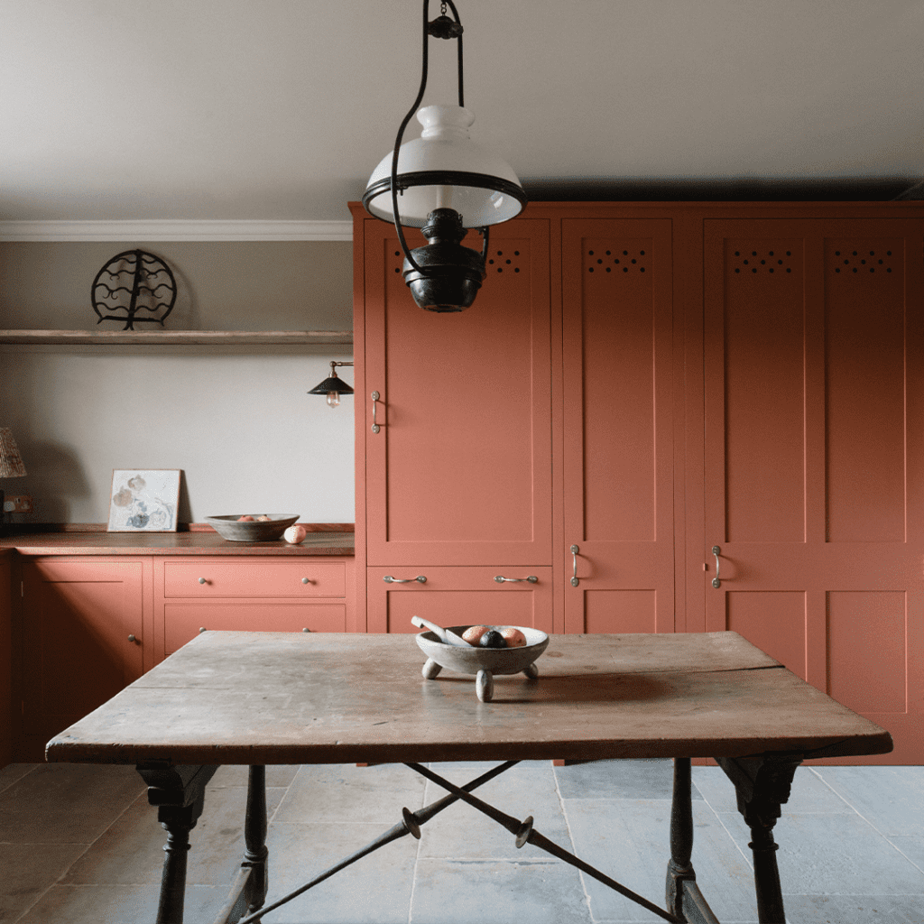 Custom cabinetry in warm terracotta, featuring tall cupboards and lower cabinets with brass handles, complemented by a rustic wooden table.