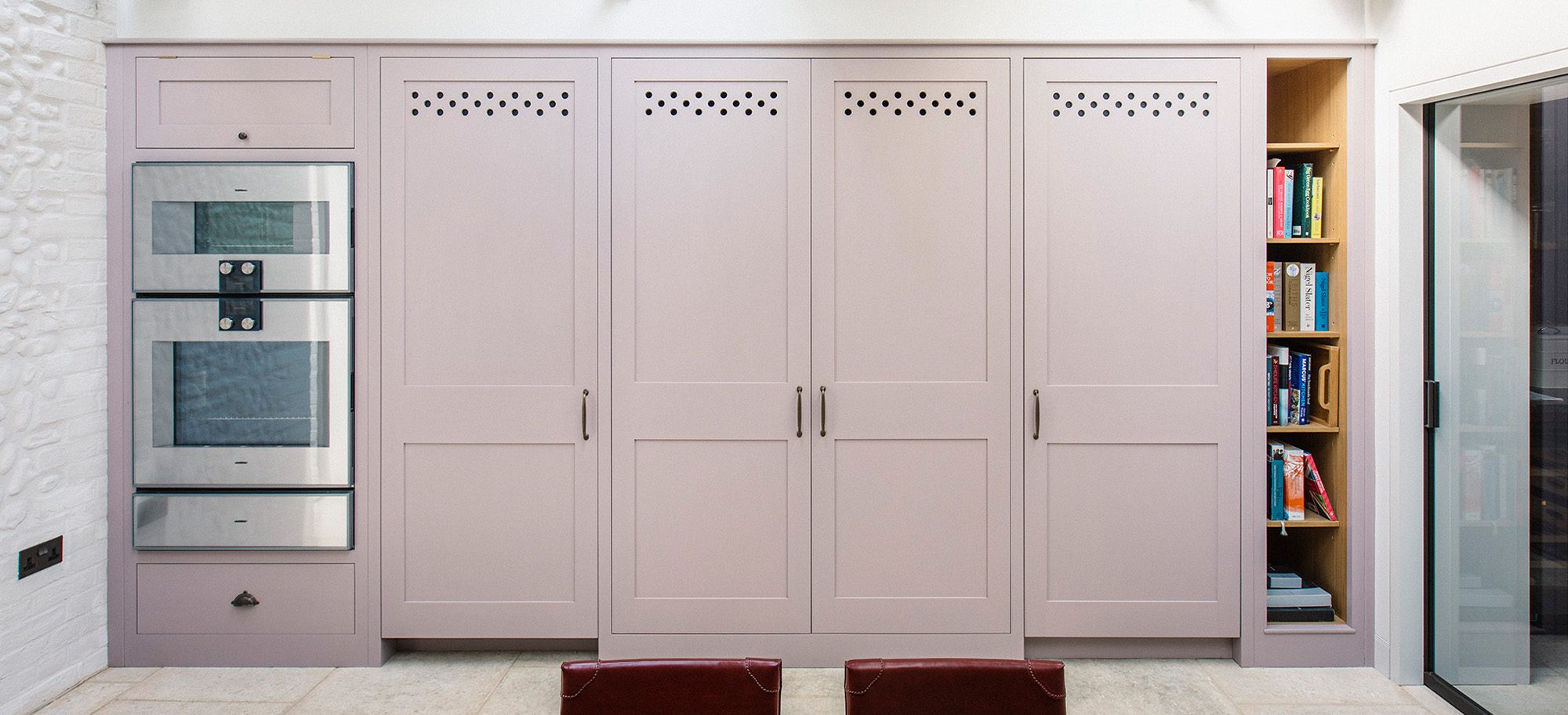  Bespoke cabinetry in soft pink with custom perforated detailing, integrated stainless steel appliances, and open shelving.