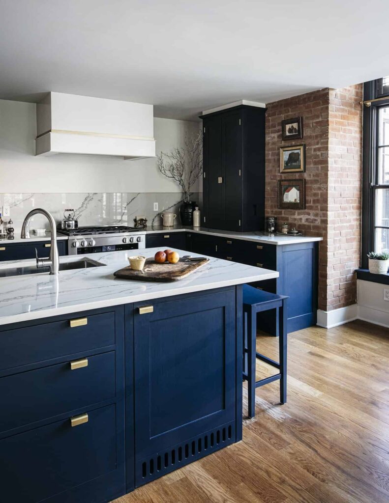 a stylish kitchen with navy blue, shaker-style cabinetry with gold-toned pulls, white marble countertops, a matching kitchen island, and exposed brick accents