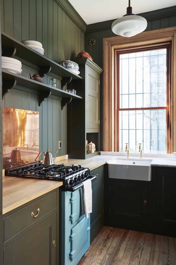a kitchen and shaker cabinetry painted in a desaturated, bold green hue with warm-toned brass fixtures and a light blue AGA range cooker