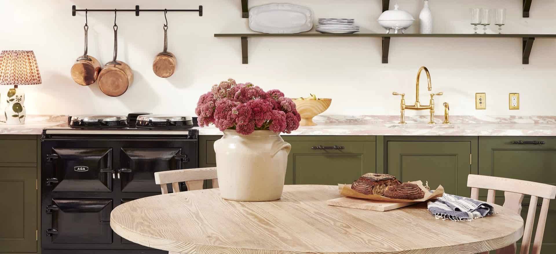  a traditional-styled kitchen with an AGA cast iron range in a black finish, with olive green cabinetry and a light wood countertop and a light wood table with a crock holding a boquet of pink flowers