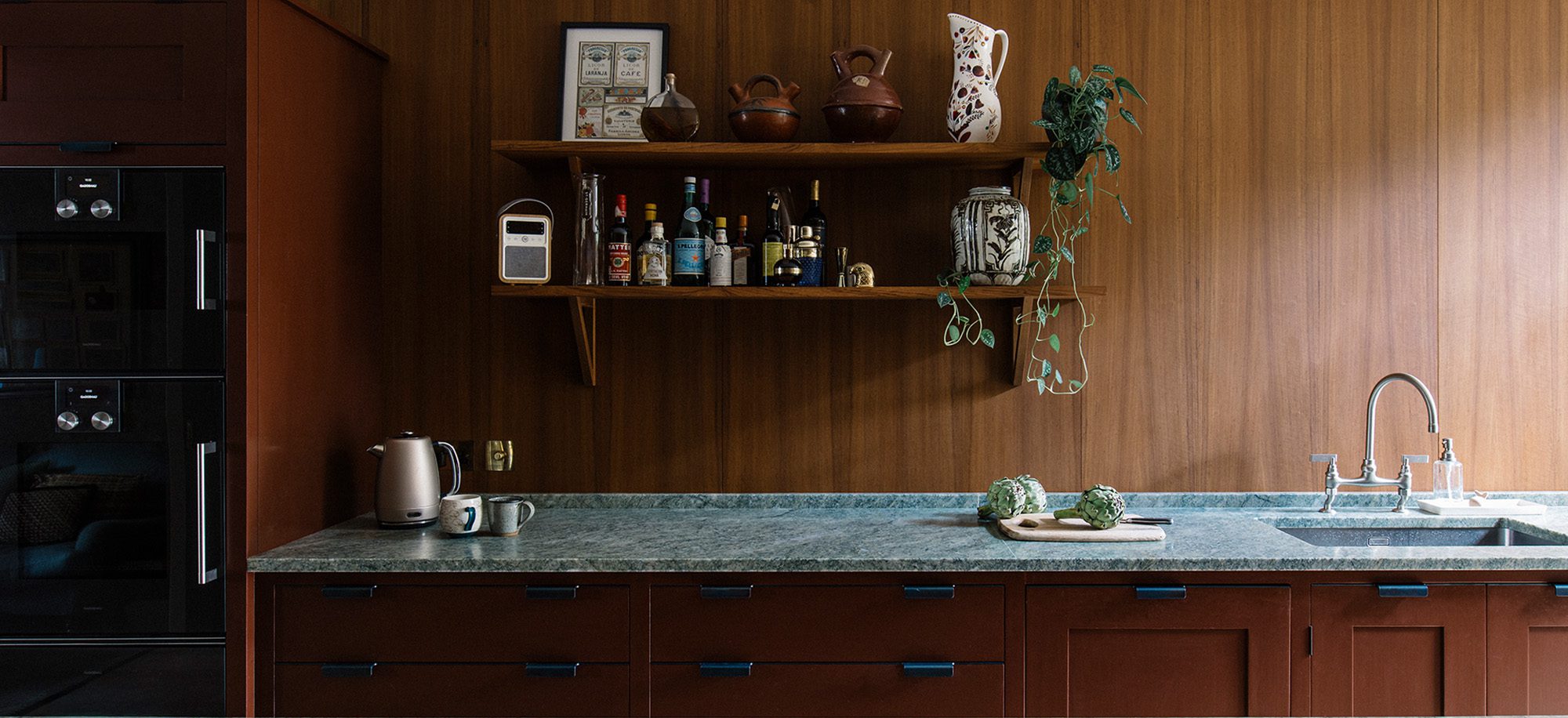  a kitchen with teak wall paneling and a green barble countertop with hand-painted open cabinetry painted in a rich chocolate tone
