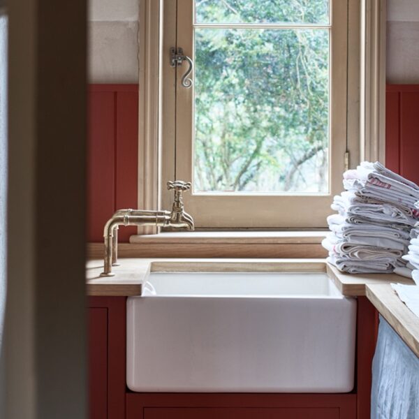 A kitchen with a farmhouse sink, brass faucet, red cabinetry, and a stack of linens by a window overlooking greenery.