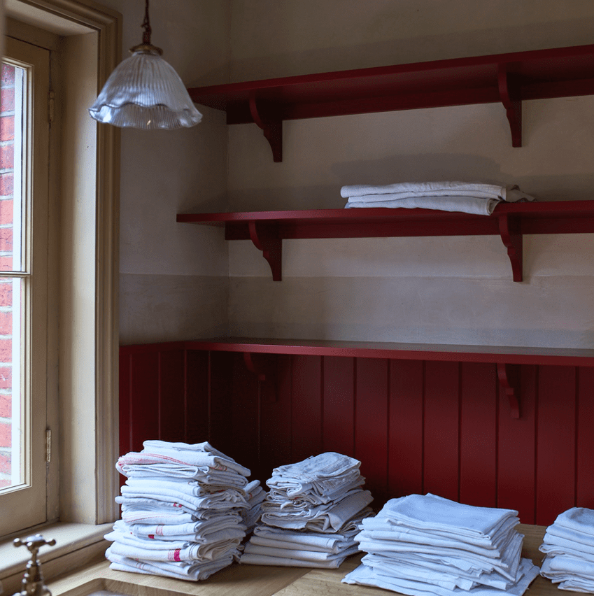 a custom utility room or laundry room with open wooden shelving painted in a rich, red color called Rusty Nail with stacks of white linens neatly folded on the counter.