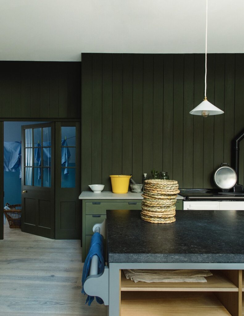 A kitchen with dark green paneled walls, a black countertop island, a yellow bucket, and a pendant light, leading to a blue room with a laundry basket.