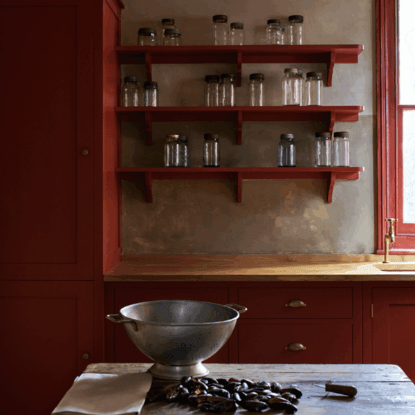 A rustic kitchen with deep red cabinetry, open shelves holding glass jars, a wooden countertop, and a metal colander on a wooden table.