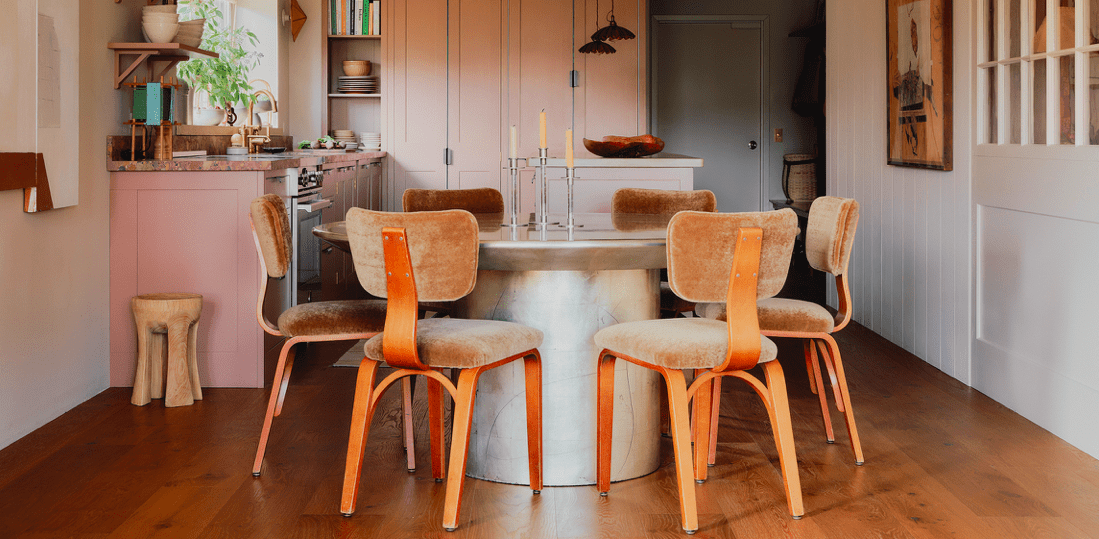  A warm, elegant kitchen features a round table with six vintage-style chairs, pink cabinetry, wooden floors, and soft lighting, creating a cozy, inviting atmosphere.